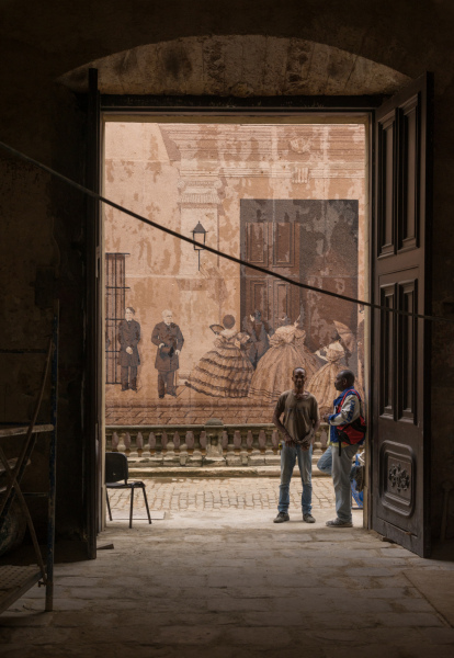 January 8, 2016 - Havana, Cuba: Two men stand in the doorway of apartment building across from a large mural depicting Cuban history. (Liz Roll/Polaris)